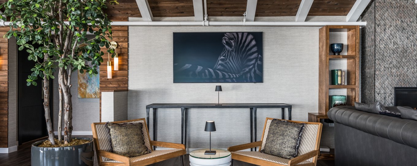 Modern living room with wooden chairs, a zebra artwork, potted tree, and tufted leather stools. Neutral tones and wood beams enhance the cozy atmosphere.